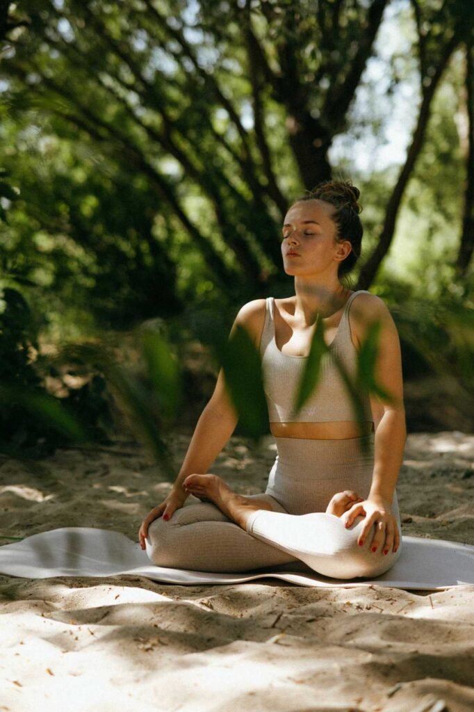 A woman practicing holistic health doing outdoor meditation, sitting cross-legged in a yoga pose on a mat in a peaceful forest setting, surrounded by natural sunlight and greenery.